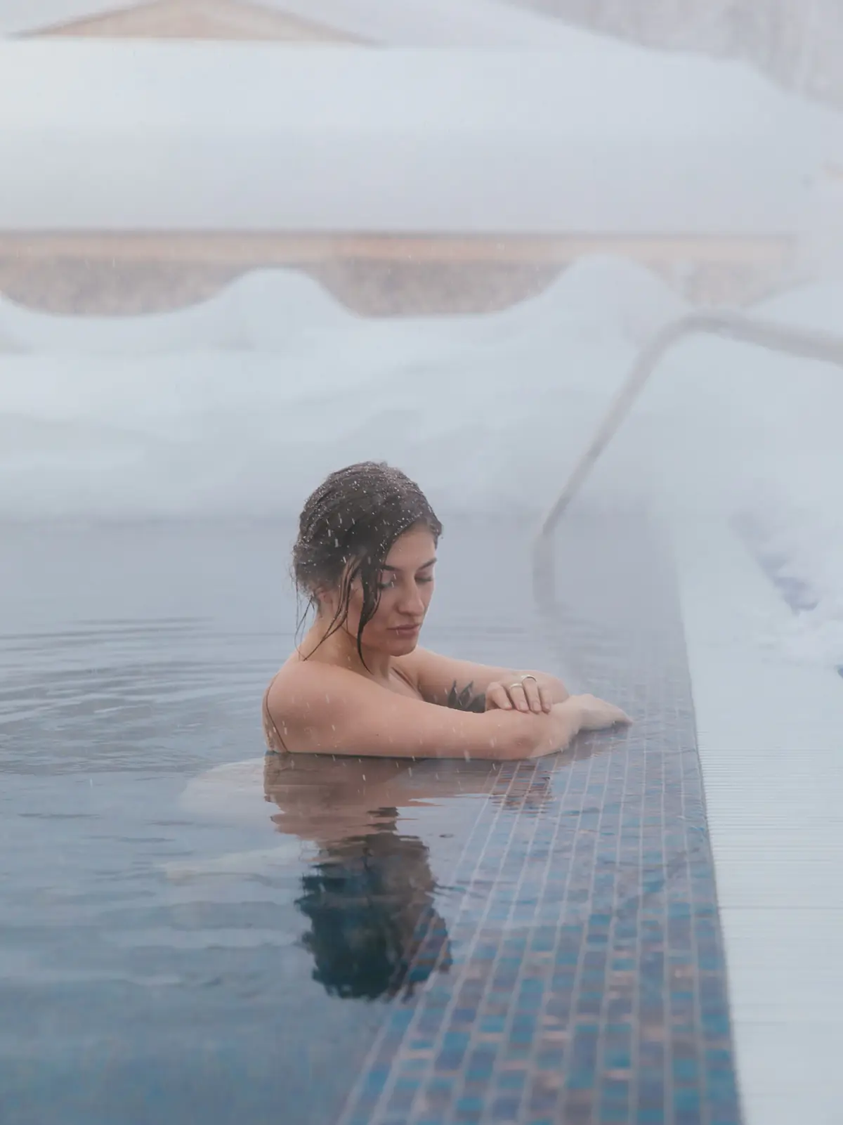 A woman resting her arms on the edge of an outdoor pool as snow falls