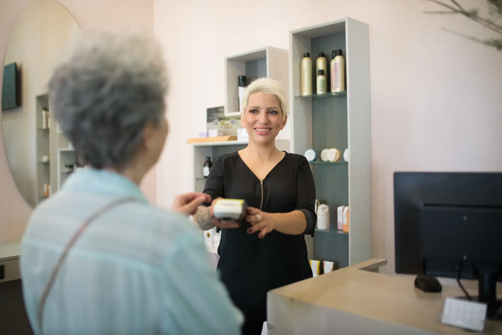 A smiling staff member holding out a card reader to a guest across a front desk, with shelves of skincare products behind
