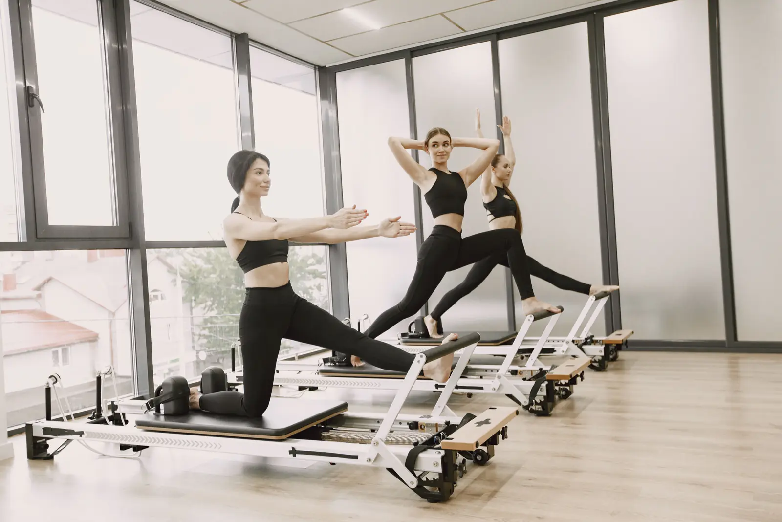 Three women doing kneeling lunges on reformers in a bright studio lined with windows