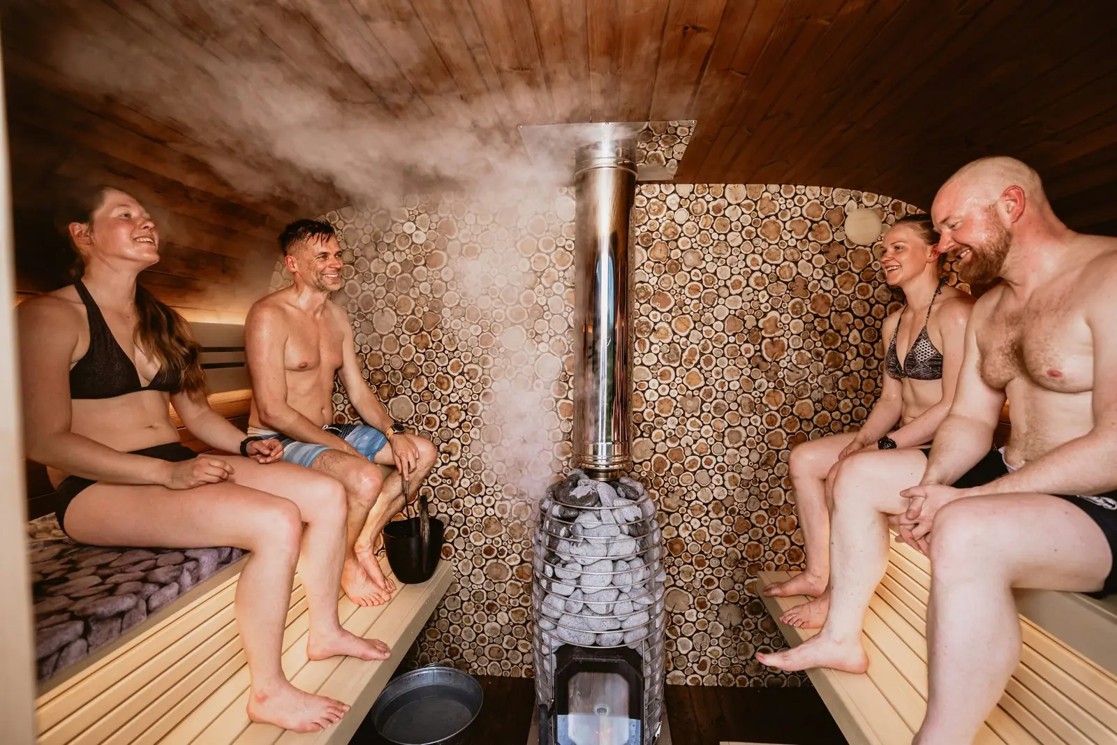 Four friends in swimwear laughing on the benches either side of a steaming stove in a barrel sauna