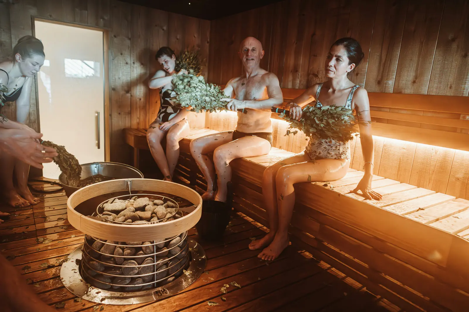 A group sharing a warmly lit wooden sauna around a central stone heater, holding leafy birch bundles