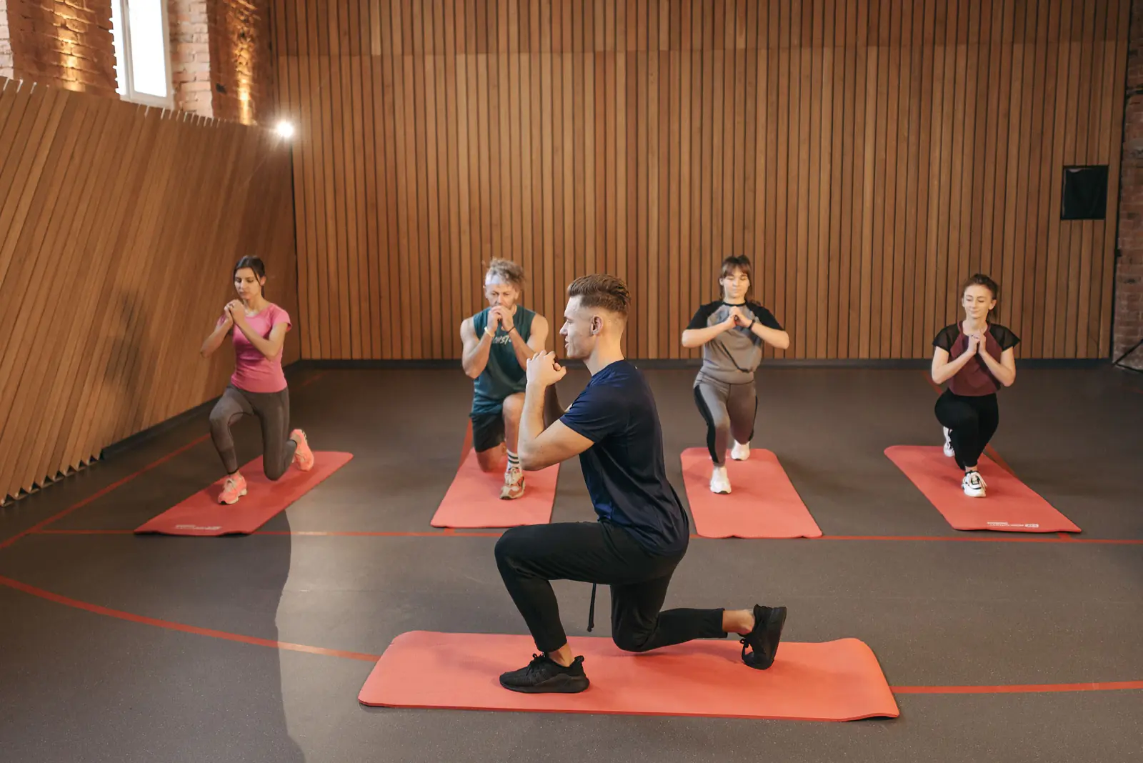 A coach demonstrating a split lunge to four members on mats in a wood-panelled studio